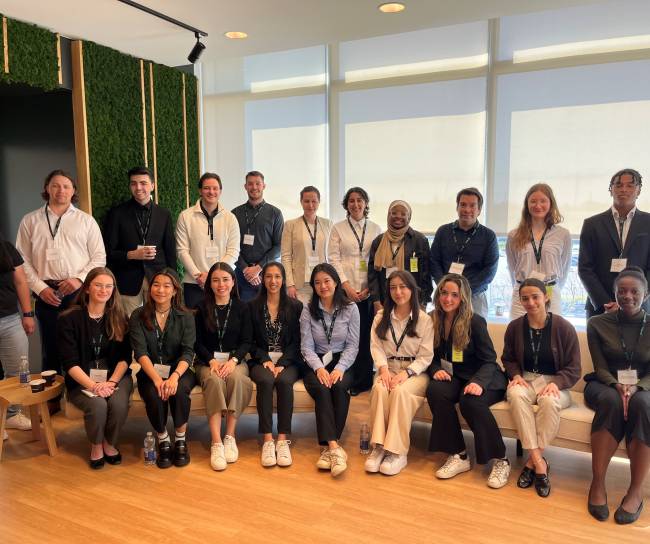 Group photo of diverse young professionals at an office event, with participants seated and standing in a well-lit modern room with wood flooring and greenery on the wall.